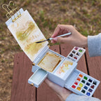 White Field Studio watercolor palette held during an outdoor sketch, with paint drawers extended beside the paper support.